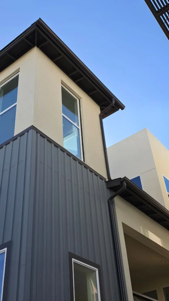 Dark brown rain gutters and matching downspouts installed on a modern two-story home with charcoal gray vertical siding, cream stucco walls, and large windows in San Diego.